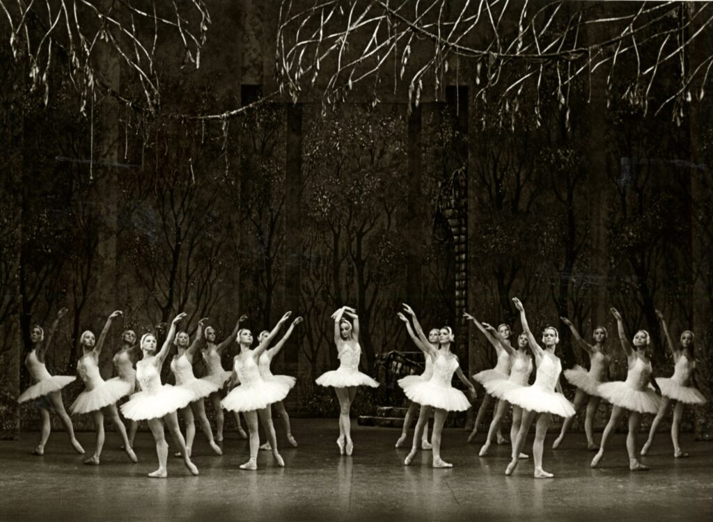 Valkoisiin tutuihin pukeutuneet naistanssijat muodostelmassa, jossa keskellä on yksi tanssija kärkitossuilla. Mustavalkoinen kuva. 