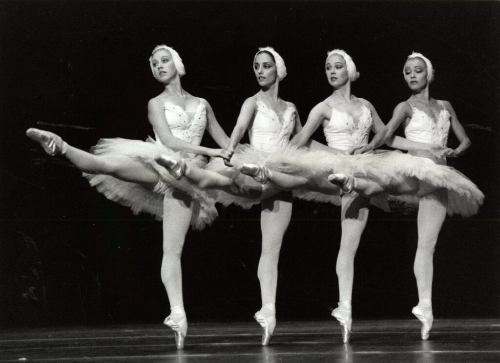 Mustavalkoinen kuva, jossa neljä valkoisiin tutuihin ja päähineisiin pukeutunutta tanssijaa identississä asennoissa. He ovat rivissä ja pitävät toisiaan käsistä kiinni. Tanssijat ovat kohottaneet oikeat jalkansa vaakasuoraan. 