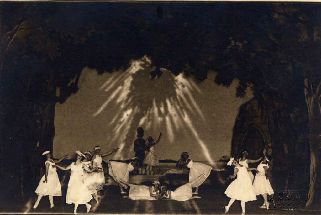 Mustavalkoinen kuva, jossa valkoasuisia balettitanssijoita erilaisissa asennoissa. Heidän keskellään seisoo pääosia esittävät nais- ja miestanssijat. 