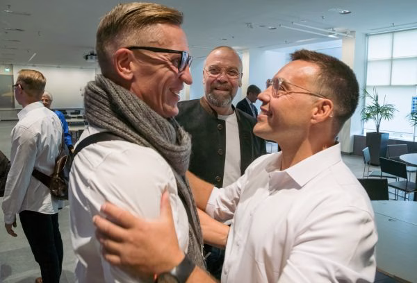 Kaksi iloiselta näyttävää henkilöä halaavat kuvassa etualalla. Heidän takanaan seisoo kolmas henkilö.