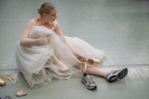 Balettitanssija istuu lattialla ja vaihtaa kärkitossujen tilalle lenkkitossut.