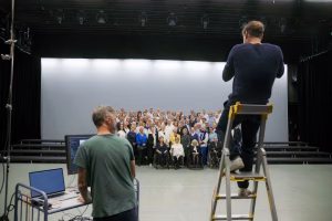 Valokuvaaja ja hänen avustajansa työskentelevät tietokoneen ja tikkaiden kanssa. Takana ihmiset ovat asettautuneet ryhmäkuvaa varten.