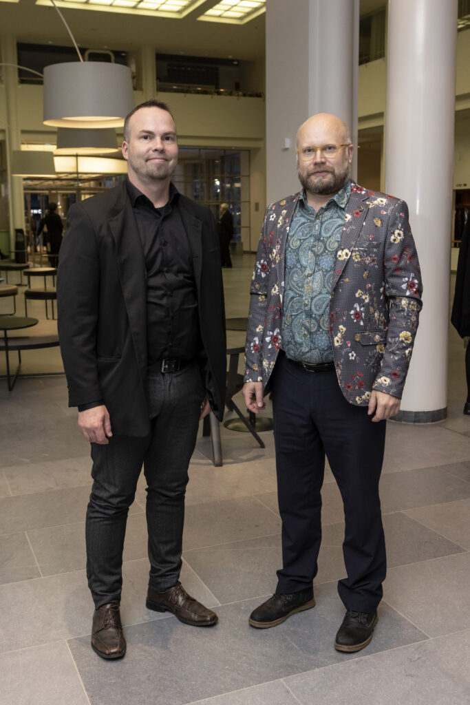 Kaksi henkilöä seisoo Oopperatalon lämpiössä pylväiden edessä. Toisella on täysin musta asu ja toisella värikäs ja kuviollinen paita ja takki.