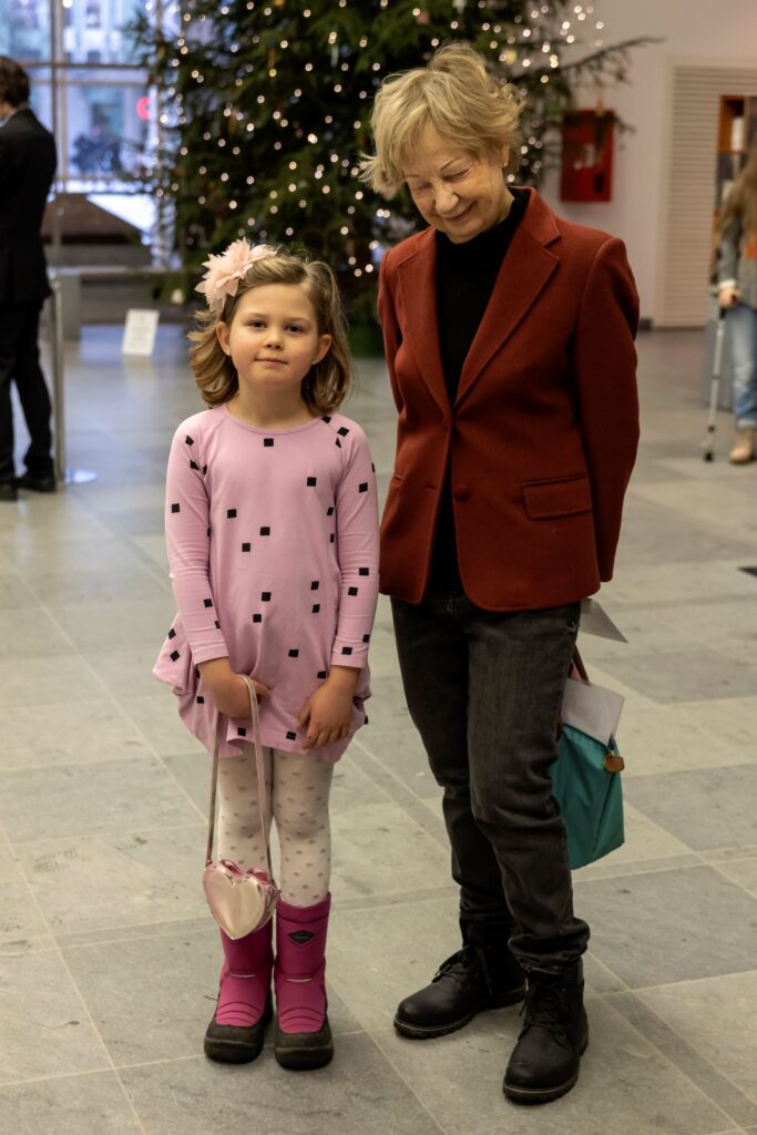 Lapsi ja aikuinen seisovat vierekkäin Oopperatalon lämpiössä koristellun joulukuusen edessä. Lapsen asu on vaaleanpunainen ja hänellä on hiuksissaan vaaleanpunainen hiuskoriste. Aikuisella henkilöllä on vaaleat, lyhyet hiukset ja tummanpunainen jakku.