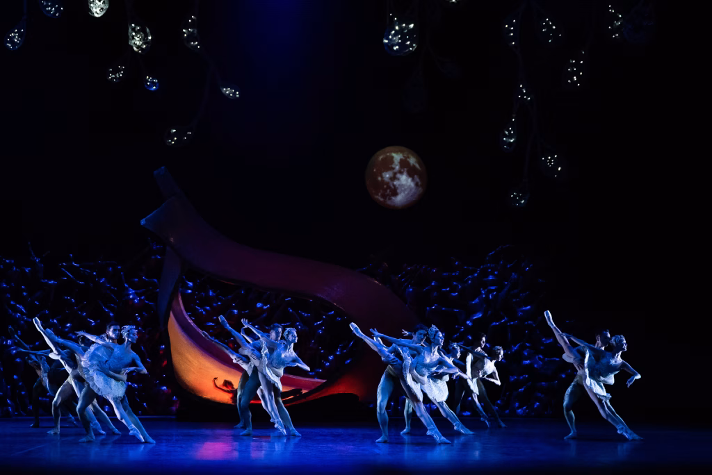 Joukko tanssijoita sinisessä valossa lavalla, taustalla kuu ja tähtiä. Takana makaa yksi tanssija oranssin, hämärästi valaistun lavasteen päällä.